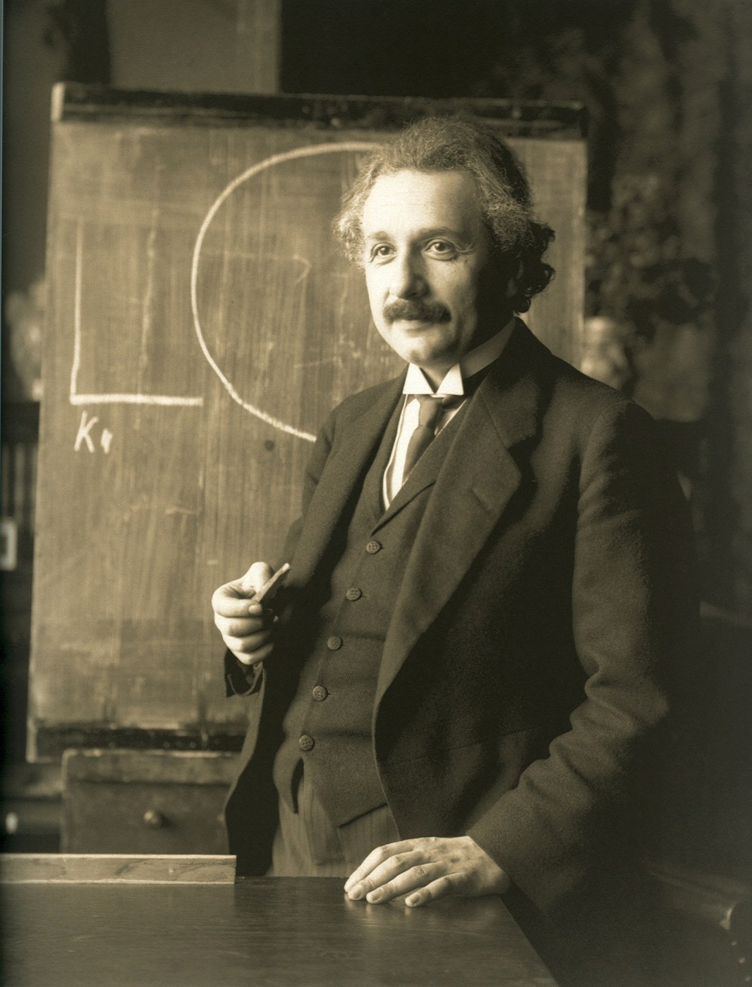 Original black-and-white photograph of Albert Einstein standing beside a blackboard holding a piece of chalk
