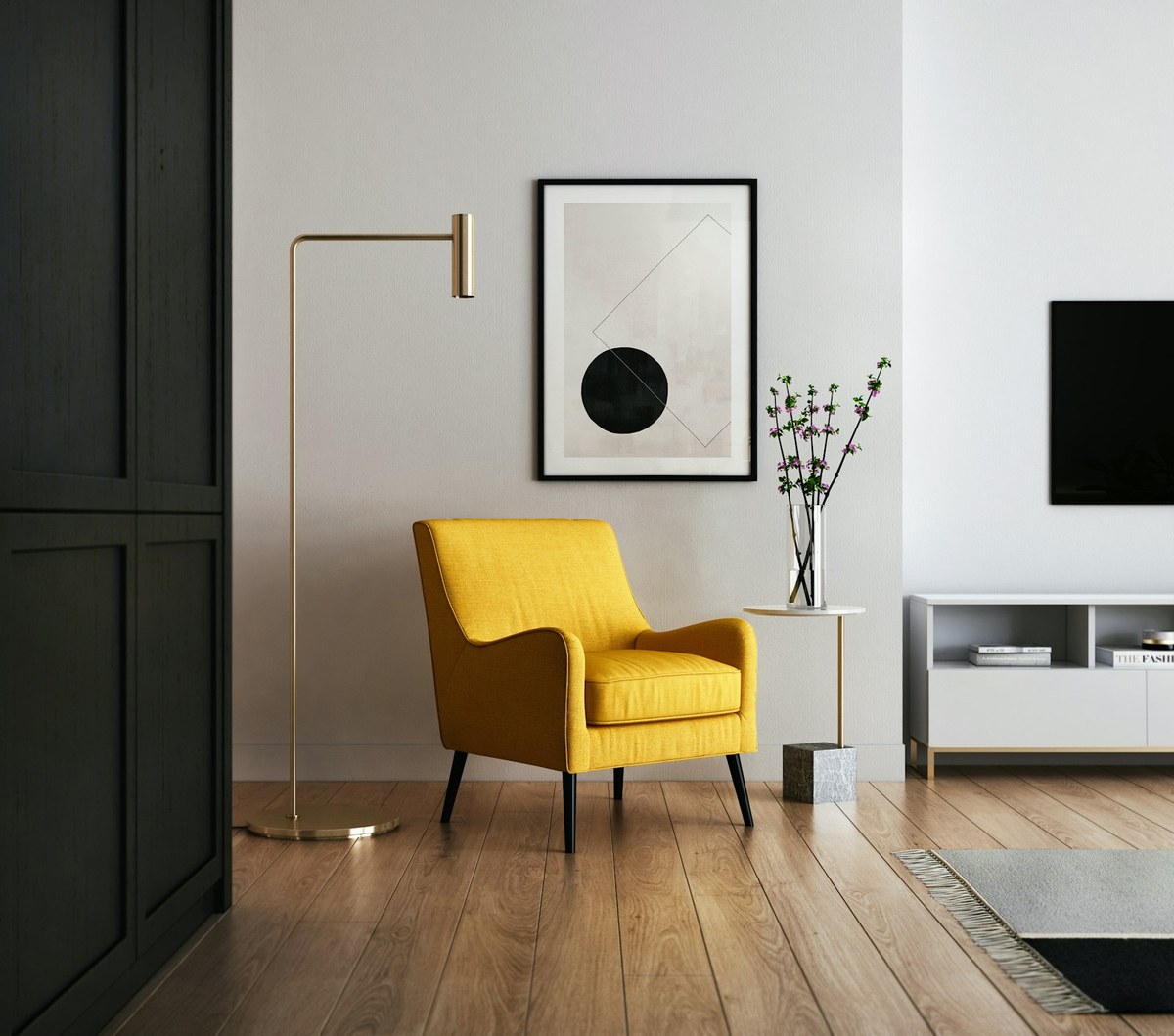 A bright living room photographed straight on: a mustard yellow armchair against an off-white wall, a brass floor lamp to its left, a framed print of a black circle and a thin diamond outline above it, a slim side table holding a glass vase of pink blossom, a dark wardrobe at the far left, a wall-mounted TV and a low white sideboard at the right, oak plank flooring